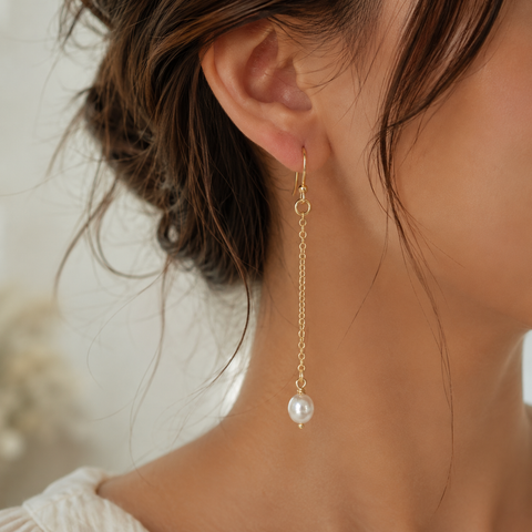A close-up of a woman's ear wearing a delicate gold dangle earring with a single white pearl at the end, set against a soft, blurred light background by Judy McPhee of Salt Spring