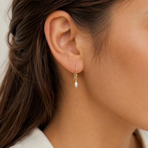 A close-up of a model's ear wearing a delicate gold dangle earring with a small white pearl drop, shown against her skin and brown hair.
