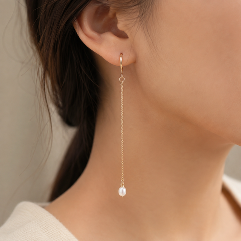 A close-up of a model's ear wearing a delicate gold dangle earring with a small white pearl at the end, set against a soft neutral background.