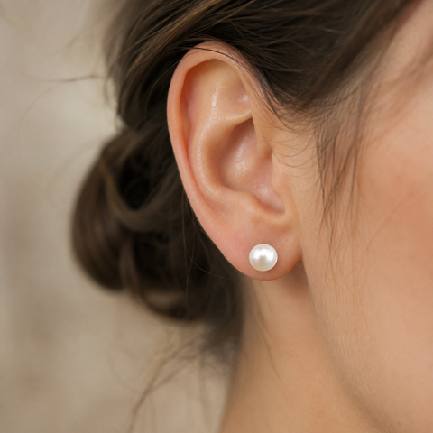 A close-up of a woman's ear wearing a single round white pearl stud earring against a soft beige background.
