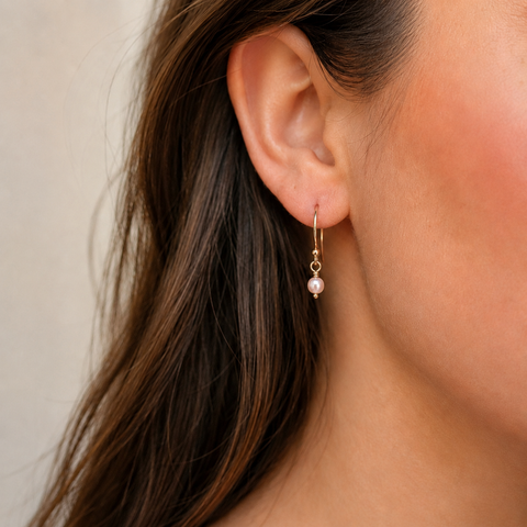 A close-up of a woman's ear wearing a delicate gold huggie hoop earring with a small white pearl drop.